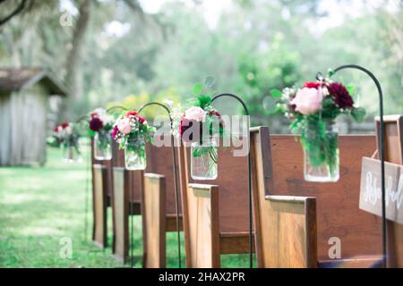 Mason jars with pink and red flowers decorations hanging at wedding ceremony isle with wood pews outdoors Stock Photo