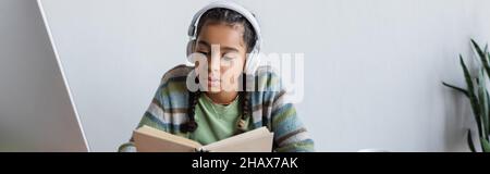 african american teenage girl in headphones reading book while studying at home, banner Stock Photo