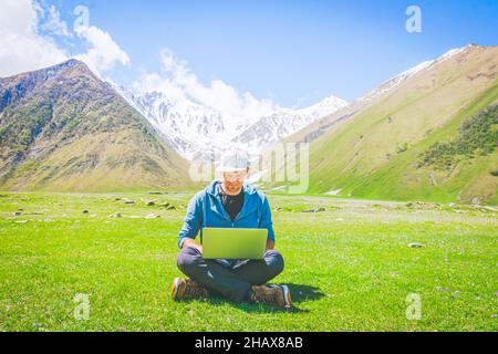 Middle age male person sits with crossed legs on the grass with laptop on his knees and types with smile in his face. Remote work and blogging while t Stock Photo