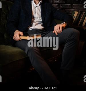 Unrecognizable stylish man looks at his watch while sitting on the couch with a book. Stock Photo