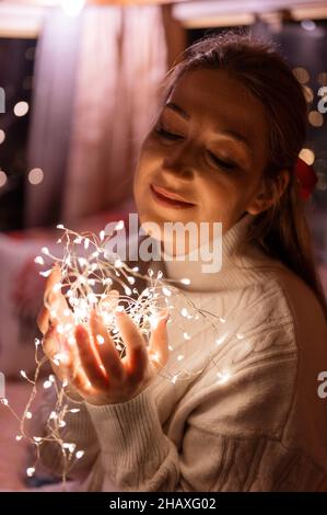 beautiful young candid woman holding in hands near the face a bright led garland of glowing lights and enjoyment an inspiring mood relaxing at home on Stock Photo