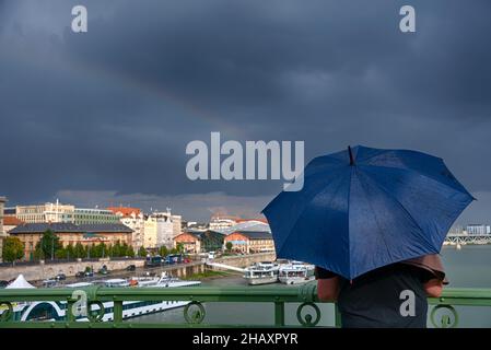 Person standing on a bridge under umbrella watches rainbow over the city. Rear view Stock Photo