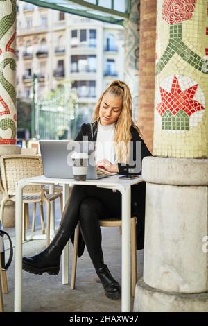 Concentrated female freelancer taking notes in planner while sitting at table with laptop and coffee and working in cafe Stock Photo