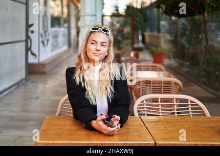 Smiling young blond female in stylish black jacket and sunglasses sitting at table in veranda of cafe Stock Photo