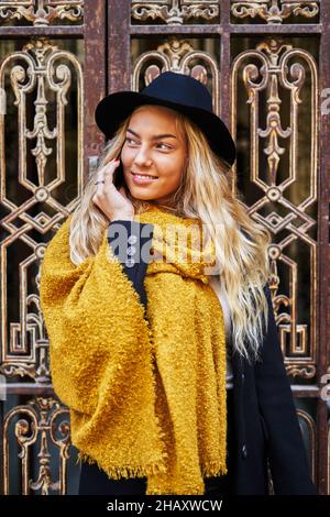 Young female in stylish black hat and woolen scarf talking on cellphone while standing at ornamental old door of building Stock Photo