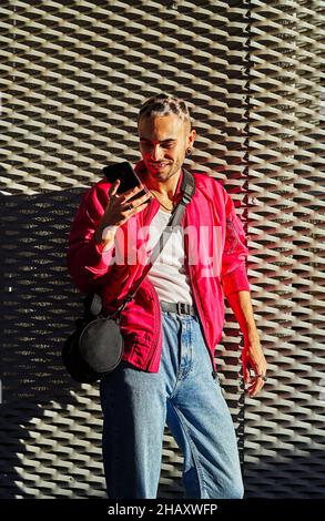 Confident eccentric male with dyed hair wearing stylish outfit standing at sunlight surfing on mobile phone Stock Photo