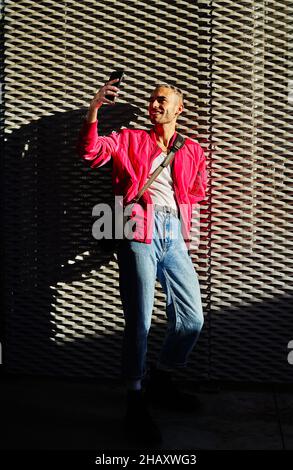 Confident eccentric male with dyed hair wearing stylish outfit standing at sunlight taking selfie on mobile phone Stock Photo