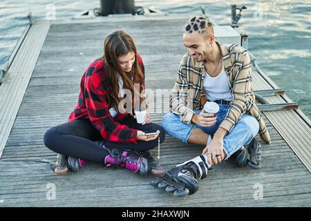 Positive eccentric man watching cellphone near female friend in casual wear and roller skates and drinking takeaway beverage on pier Stock Photo