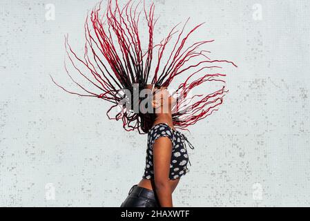 Side view of stylish African American Woman smiling while flipping long black and red hair against white wall Stock Photo