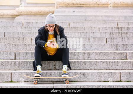 Full body of young female skater in casual clothes sitting on concrete steps of staircase of building and browsing smartphone with legs on skateboard Stock Photo