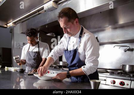 Focused professional chef wiping plate while working with black colleague pouring sauce into ceramic pot in modern kitchen Stock Photo