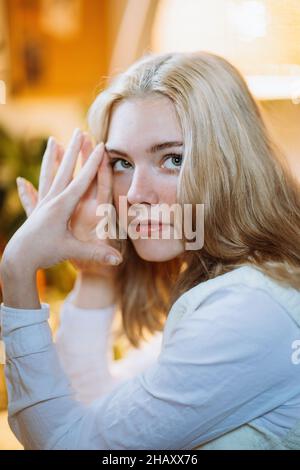 Positive young female in white blouse sitting at table touching face with hands Stock Photo