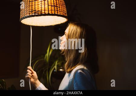 Side view of young female standing under illuminated lamp of chandelier looking at the light Stock Photo