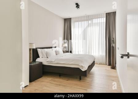 Interior of light modern bedroom with soft white bed with pillows and blanket placed near bedside table with lamp near window with curtains Stock Photo