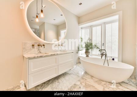 Interior of modern bathroom with marble tiled floor and white bathtub oval mirror hanging on wall above double washbasin with cabinets and bronze vint Stock Photo