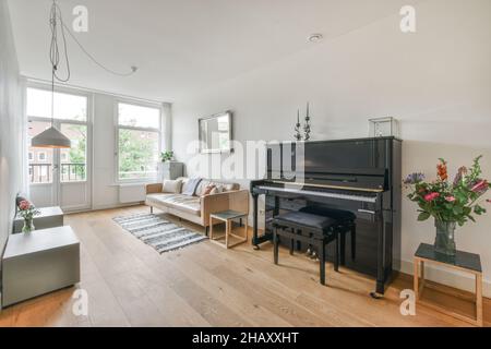 Interior of spacious living room furnished with comfortable couch and tables decorated with vase of flowers on parquet floor near big black piano agai Stock Photo