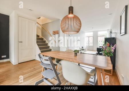 Spacious light dining zone in modern house furnished with wooden table and chairs next to stairs and open plan living room with white walls Stock Photo