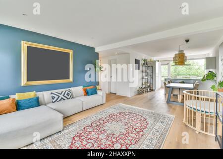 Comfortable sofa with cushions placed near windows at wall with frame in spacious living room with baby crib and dining table in apartment Stock Photo