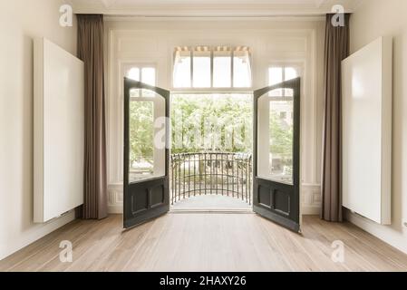 Interior of spacious empty room with white walls and laminated floor and brown curtains on open windows with a terrace in the background Stock Photo