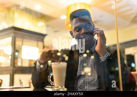 Through windows of confident young African American male entrepreneur in elegant outfit drinking glass of latte macchiato and on a phone call on smart Stock Photo