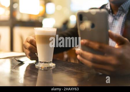 Cropped unrecognizable African American male entrepreneur in elegant outfit drinking glass of latte macchiato and messaging on mobile phone during bre Stock Photo