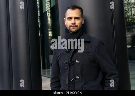 Serious male in black coat standing on street near column of contemporary building and looking at camera in daytime Stock Photo