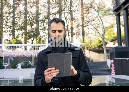 Positive male entrepreneur in black coat standing and using tablet on city street against trees in daytime Stock Photo