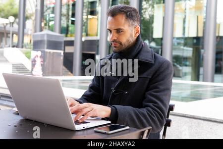 Smiling businessman in black coat browsing on laptop while sitting at table in cafe in daytime Stock Photo