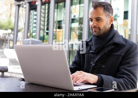 Smiling businessman in black coat browsing on laptop while sitting at table in cafe in daytime Stock Photo