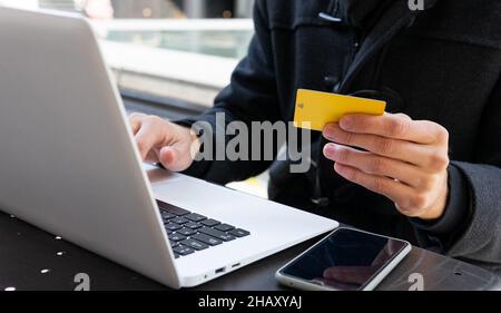 Crop unrecognizable male entrepreneur in black coat looking at details of credit card in hands while sitting at table with laptop on terrace of cafe i Stock Photo