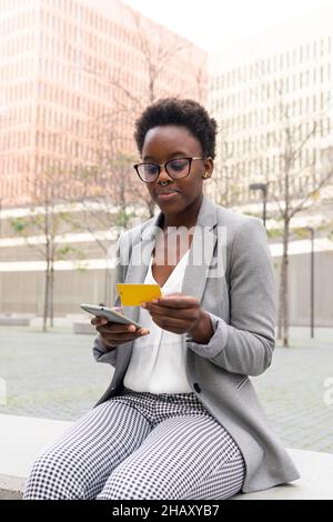 Smiling African American female entrepreneur wearing eyeglasses and jacket entering data of credit card on mobile phone during online shopping Stock Photo