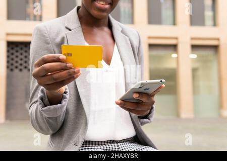 Cropped unrecognizable African American female entrepreneur wearing eyeglasses and jacket entering data of credit card on mobile phone during online s Stock Photo