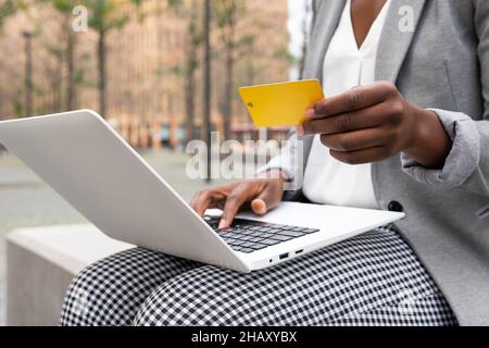 Crop anonymous black Woman freelancer sitting on terrace and ordering in internet shop on laptop and paying with plastic card Stock Photo