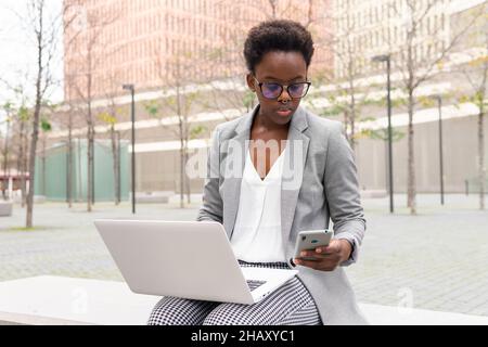 Focused young African American female freelancer in formal clothes checking notification on smartphone while working on laptop remotely sitting on ben Stock Photo
