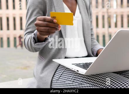 Crop anonymous black Woman freelancer sitting on terrace and ordering in internet shop on laptop and paying with plastic card Stock Photo
