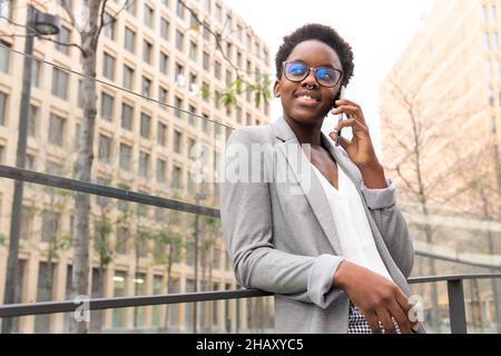 From below smiling black Woman manager wearing classy outfit browsing smartphone while standing with eyes closed on balcony with glass fence in office Stock Photo
