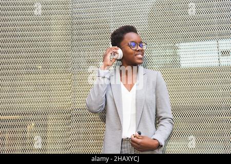 Young smiling African American female wearing elegant outfit and eyeglasses listening to music in wireless headphones while standing against metal gri Stock Photo