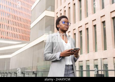Young serious African American female in eyeglasses and classy outfit browsing on smartphone walking near office building Stock Photo