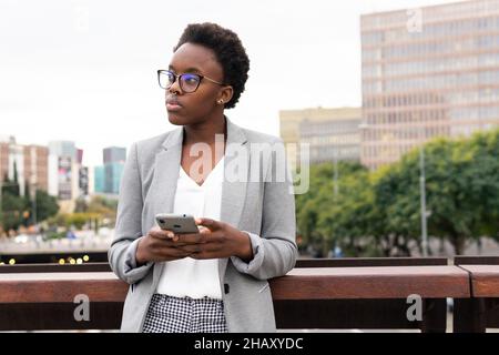 Young black female employee wearing eyeglasses and formal jacket browsing cellphone while standing on balcony of business center Stock Photo