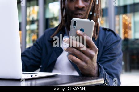 Positive African American young man messaging on smartphone sitting at table with a laptop on terrace of shopping center Stock Photo