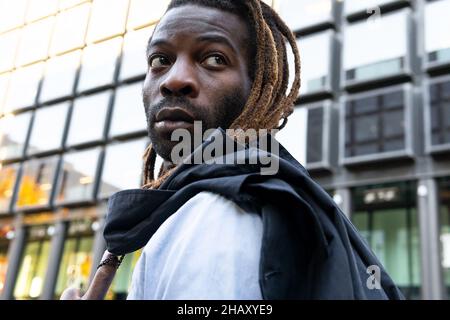 Young bearded black male with dreadlocks wearing white shirt holding the jacket over the shoulder while looking away in modern office building Stock Photo