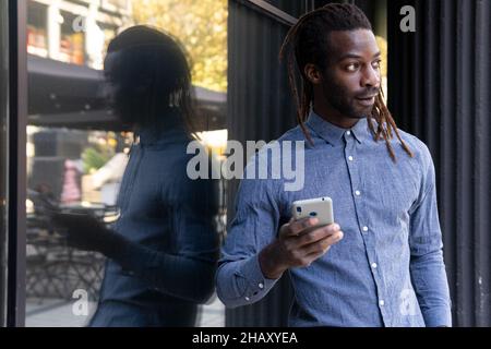 Confident African American male in casual denim shirt browsing on cellphone near window of building in daytime Stock Photo
