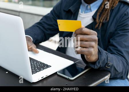Anonymous African American young man paying order in internet shop with credit card on laptop sitting at table with smartphone on terrace of shopping Stock Photo