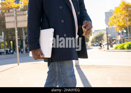 Anonymous African American male entrepreneur in navy blue jacket with laptop in street in daylight Stock Photo