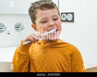Cute boy in casual wear brushing teeth while standing near white sink in bathroom during morning hygienic routine at home Stock Photo