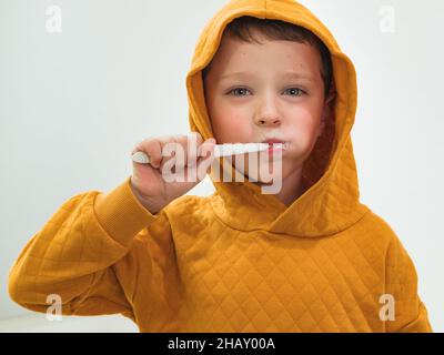 Cute boy in hoodie pullover brushing teeth in bathroom during morning hygienic routine at home looking at camera Stock Photo