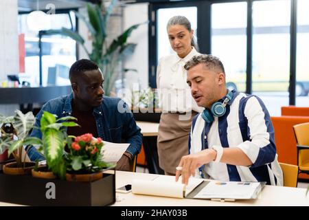 Group of multiethnic coworkers discussing business project together while doing paperwork at table with documents during work in modern office Stock Photo