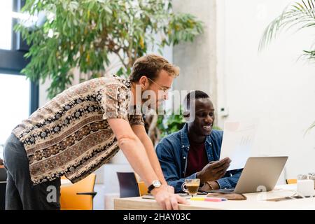 Diverse coworkers examining documents at table with laptops and green potted plants while doing paperwork together in modern light office Stock Photo