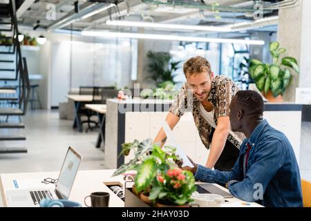 Diverse coworkers examining documents at table with laptops and green potted plants while doing paperwork together in modern light office Stock Photo
