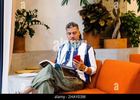 Focused male worker with headphones reading magazine and browsing on smartphone while sitting on armchair in modern office during work Stock Photo